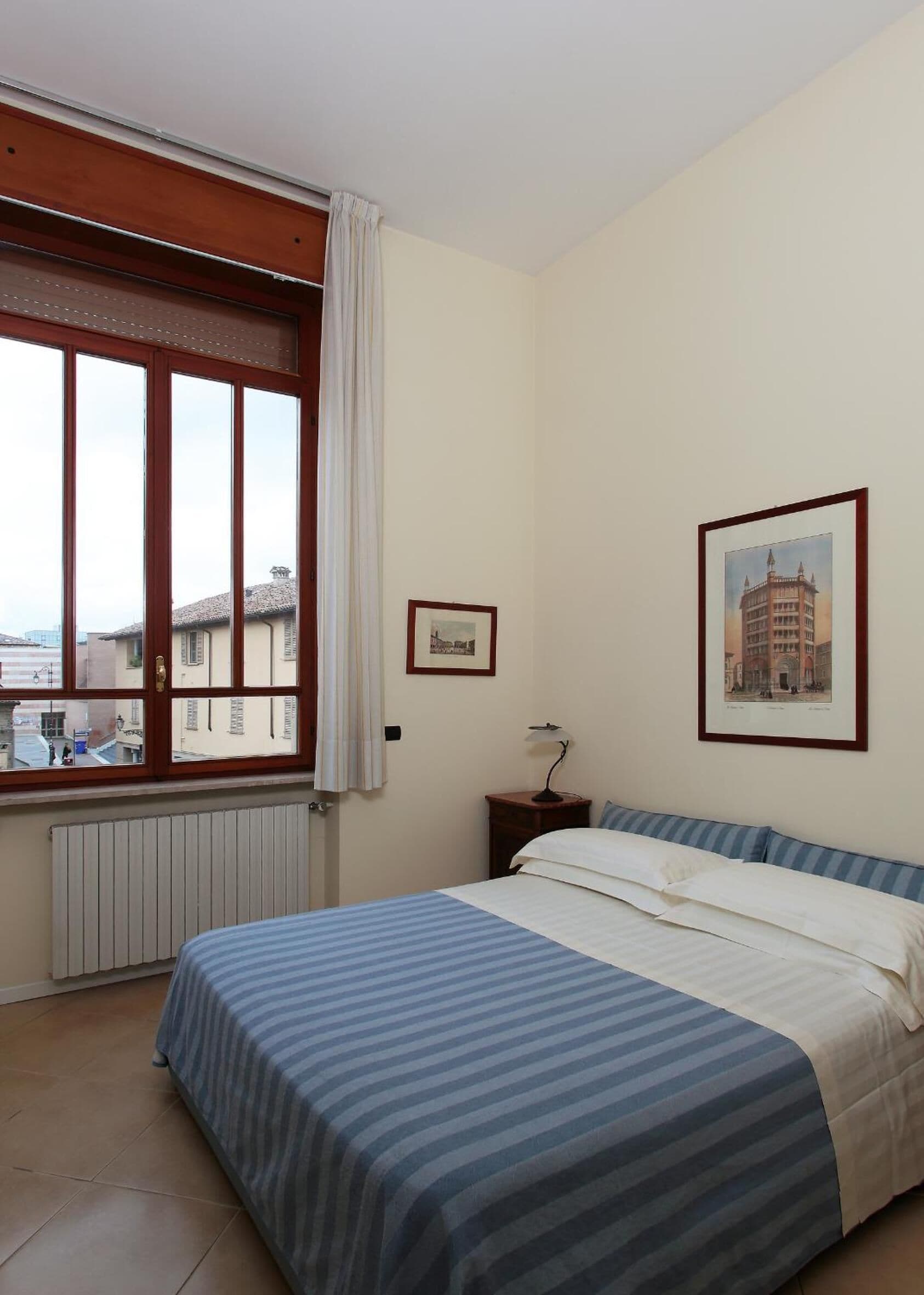 A bedroom with a large bed, a window with curtains, a radiator, a nightstand with a lamp, and two framed pictures on the wall.
