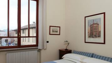 A bedroom with a large bed, a window with curtains, a radiator, a nightstand with a lamp, and two framed pictures on the wall.