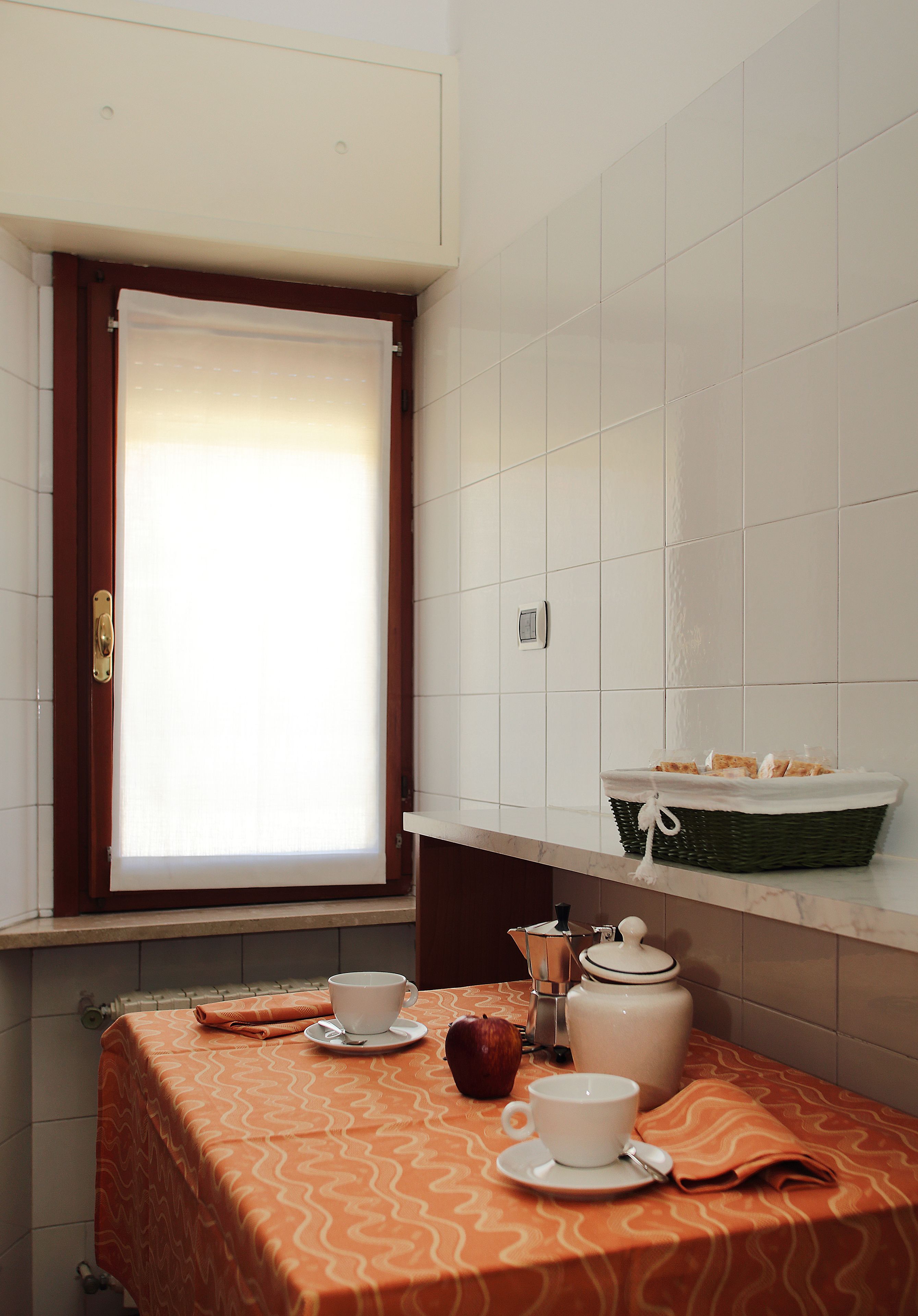 A small kitchen area with a window, a table with a patterned tablecloth, a basket, a kettle, and a cup.