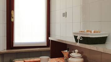 A small kitchen area with a window, a table with a patterned tablecloth, a basket, a kettle, and a cup.