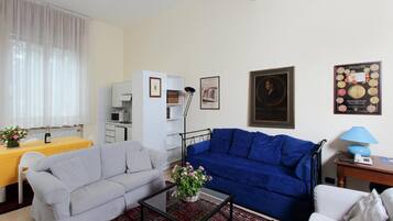Un soggiorno con un divano grigio, un divano blu, un tavolino da caffè con fiori, un tavolo da pranzo con una tovaglia gialla e un angolo cottura sullo sfondo.