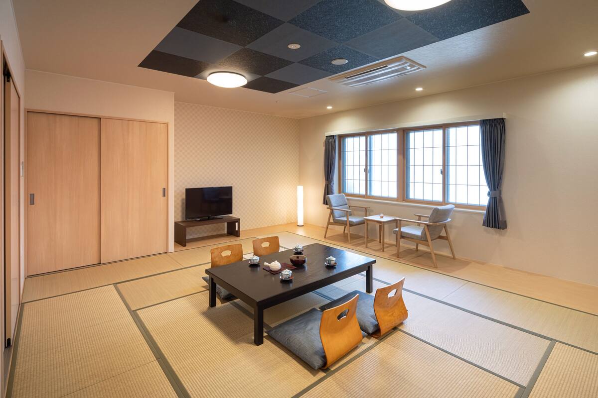 A traditional Japanese-style room with tatami flooring, a low table with cushions, a television, and a window with curtains.