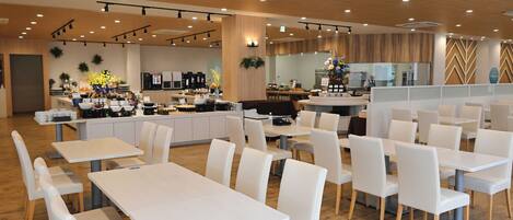 Un restaurante con mesas y sillas blancas, una zona de cocina y un mostrador de buffet.
