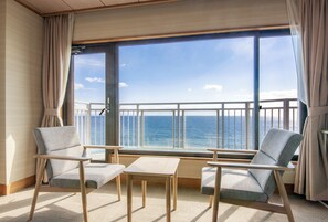 Habitación con dos sillas y una mesita pequeña, ventanas amplias con vista al mar y un balcón con barandilla.