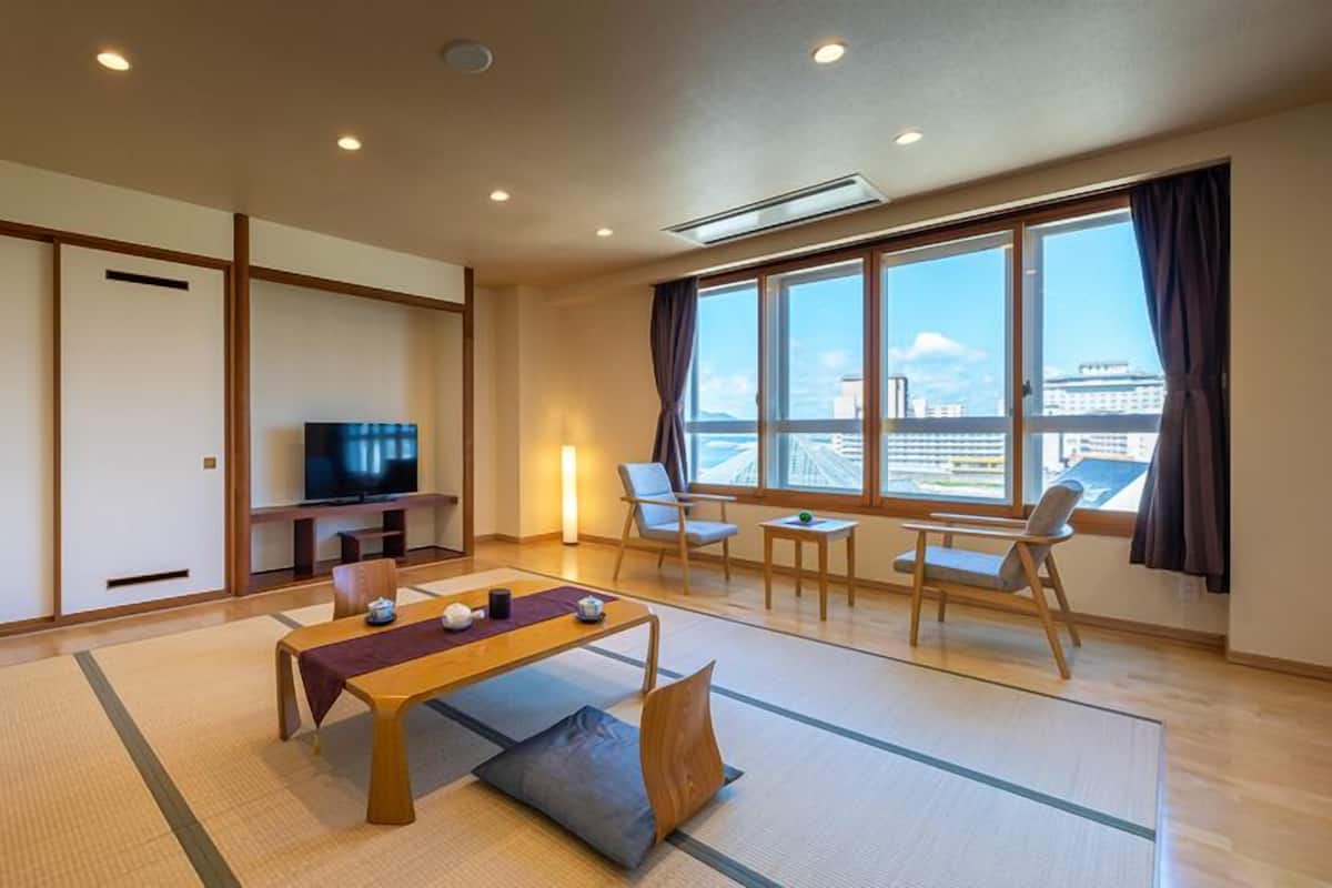 A modern hotel room with tatami mats, a TV, a sofa, a table, and large windows with a city view.
