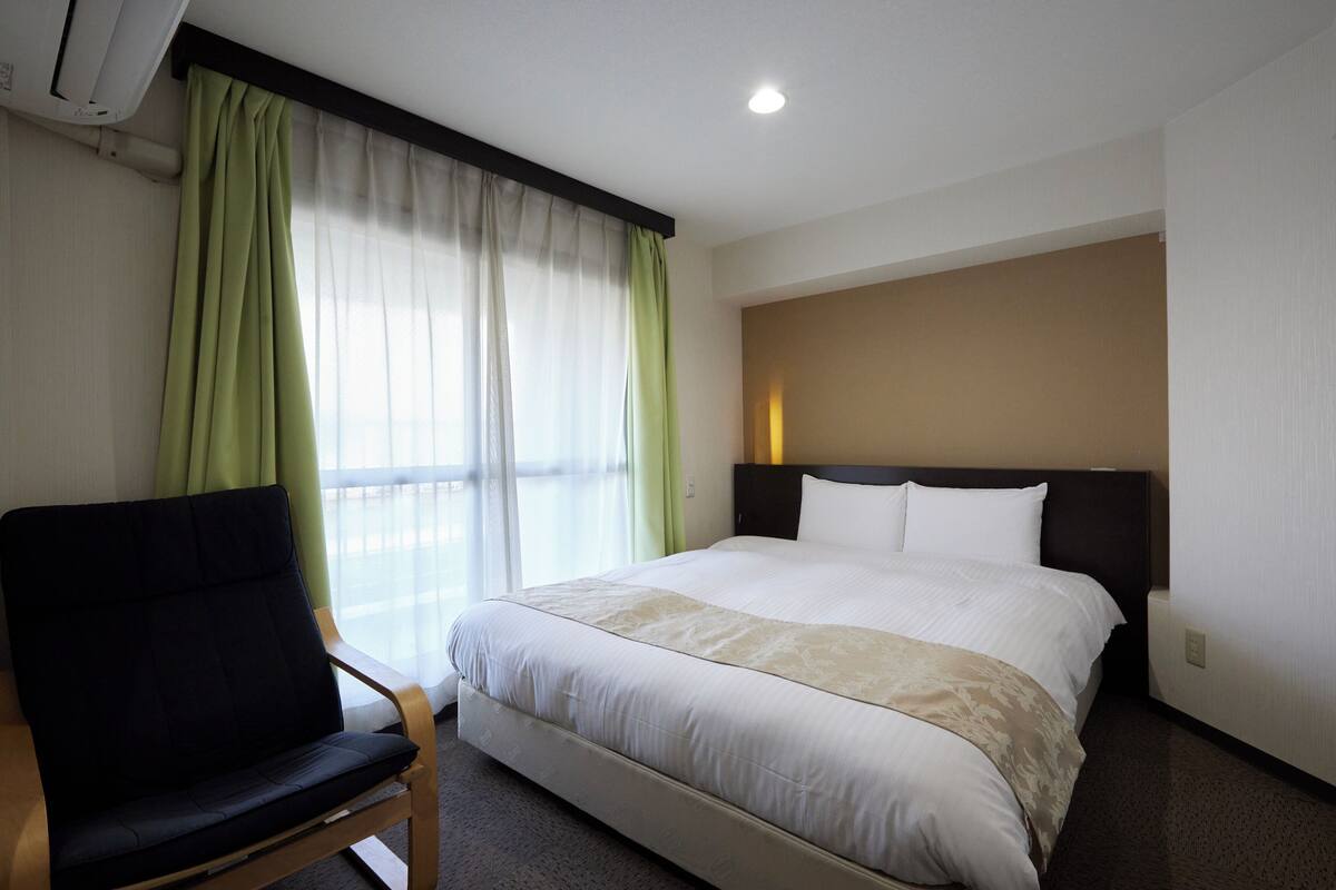 Una habitación de hotel con cama, una silla, una ventana con cortinas y un aire acondicionado de pared.
