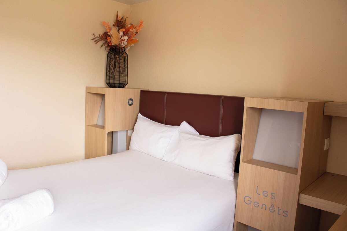 A neatly made bed with white linens, a wooden headboard, and a vase with dried flowers on the nightstand.