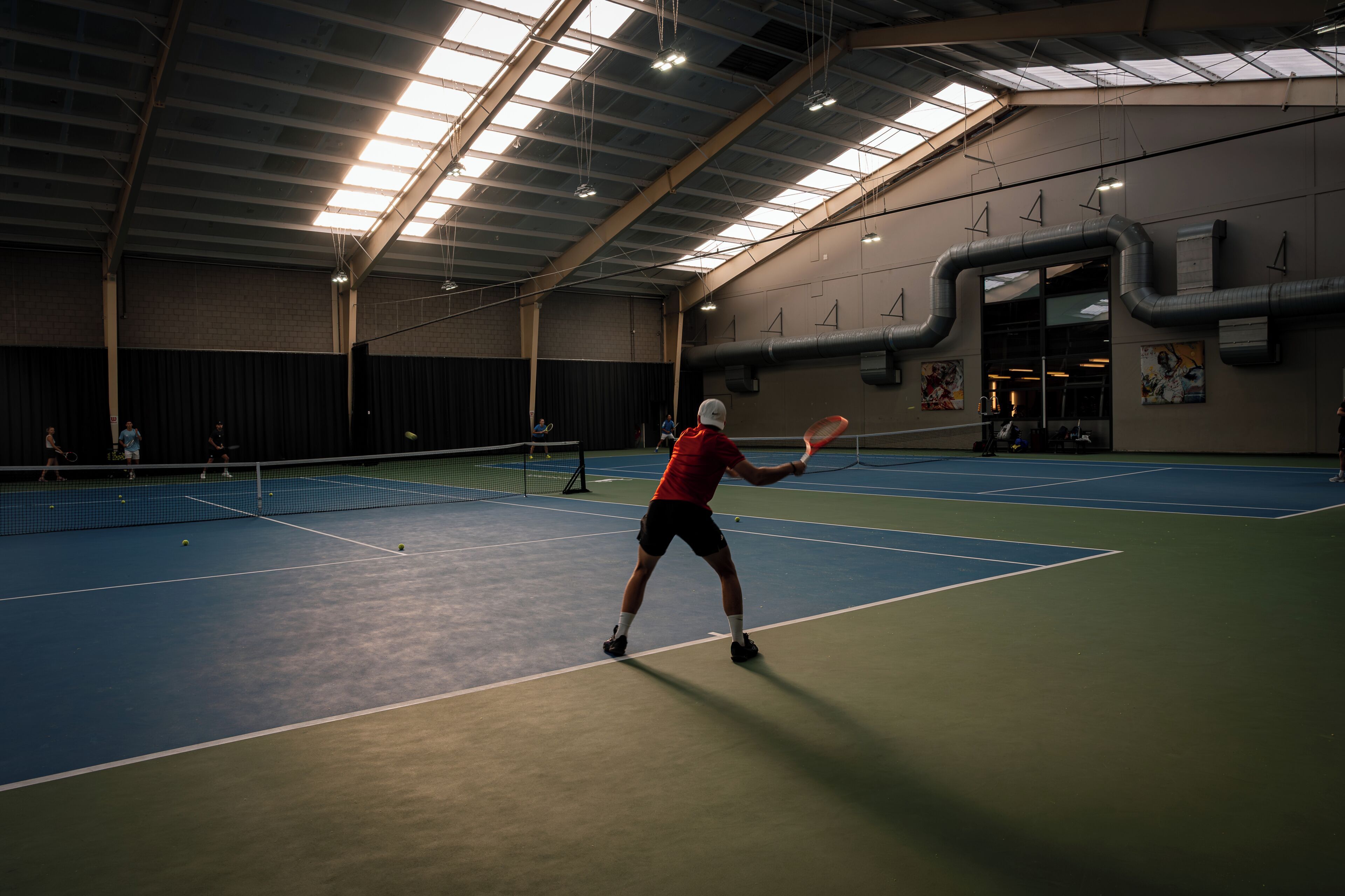 Pista de tenis interior con techo alto y tragaluces, rodeada de paredes con obras de arte.