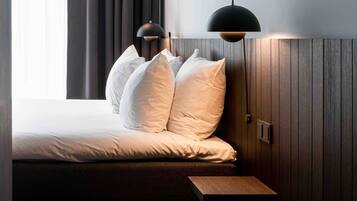 Cama bem arrumada com lençóis brancos e várias almofadas, mesa de cabeceira de madeira e duas luminárias pendentes.