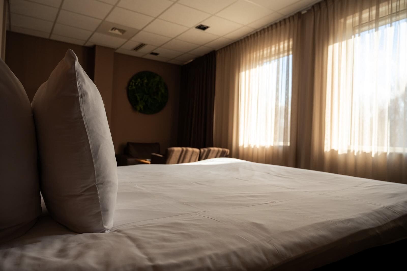 Una habitación de hotel con una cama grande, una silla, un adorno circular en la pared y una ventana con cortinas.
