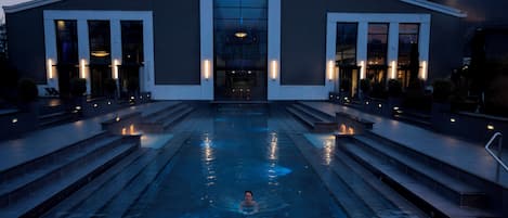 A modern building with a swimming pool in front, steps leading up to the entrance, and exterior lighting.