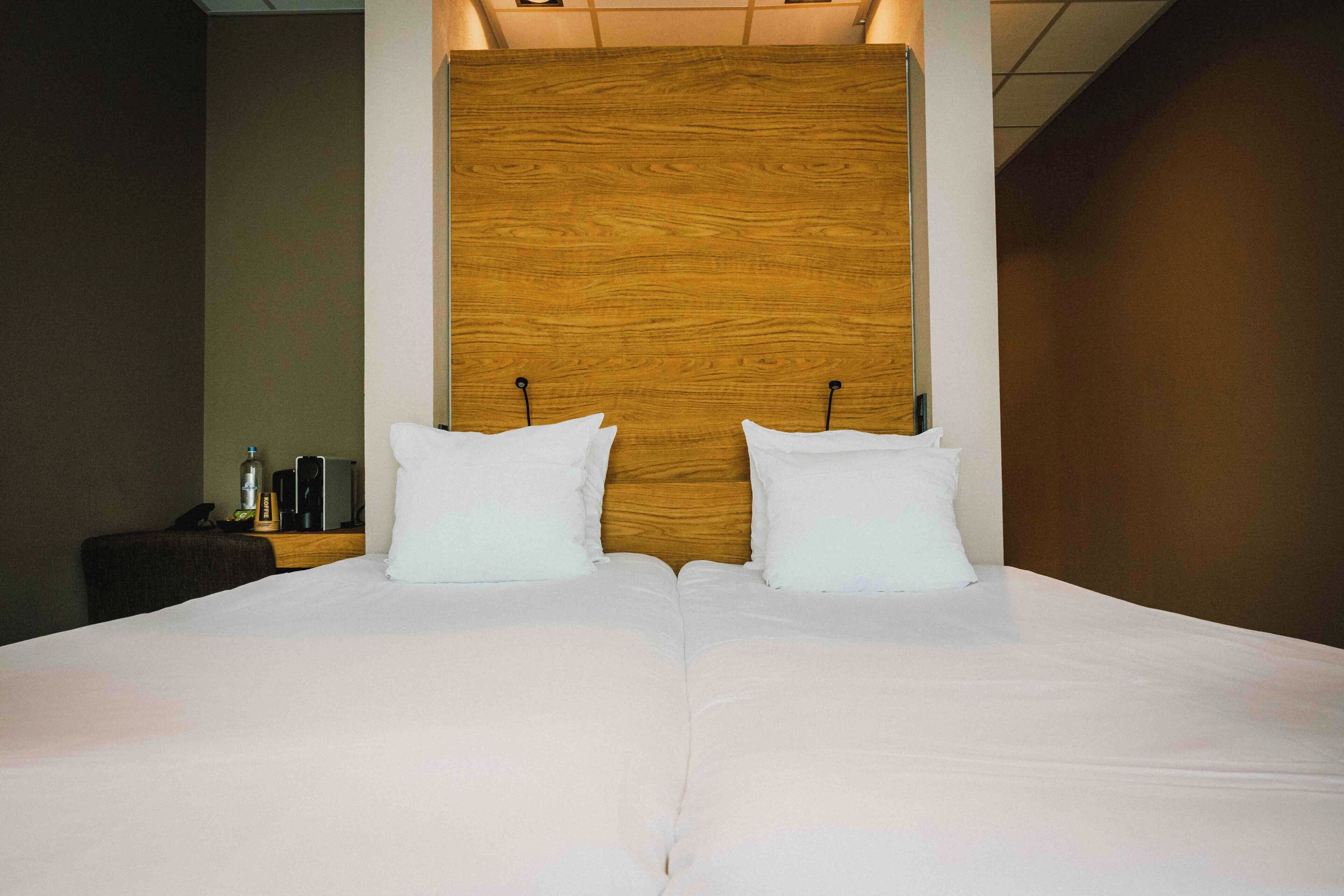 Una cama grande con ropa de cama blanca y dos almohadas, un cabecero de madera y una mesita de noche con una botella y un teléfono.