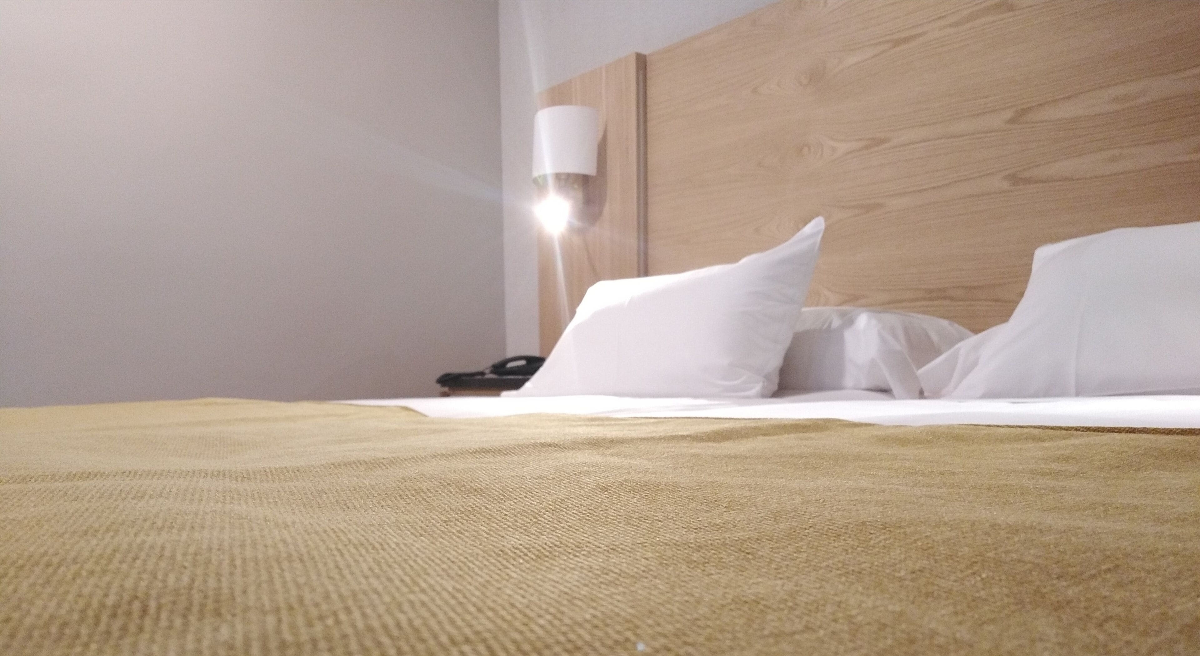 Una cama bien hecha con sábanas blancas y una manta color beige, un cabecero de madera y una lámpara de noche.
