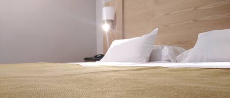 Una cama bien hecha con sábanas blancas y una manta color beige, un cabecero de madera y una lámpara de noche.