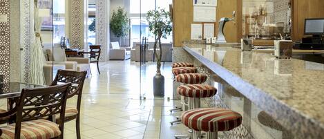 A hotel lobby with a reception desk, bar area, seating with striped stools, and a large potted plant.