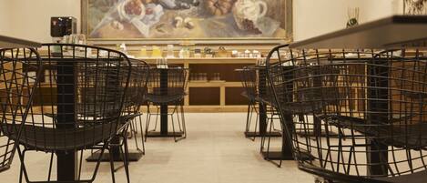 Comedor con sillas y mesas de estructura metálica, un cuadro en la pared y una zona de bar al fondo.