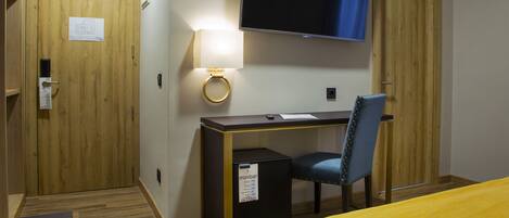 A hotel room with a wooden desk, a chair, a bed with a yellow bedspread, a television mounted on the wall, and a minibar.
