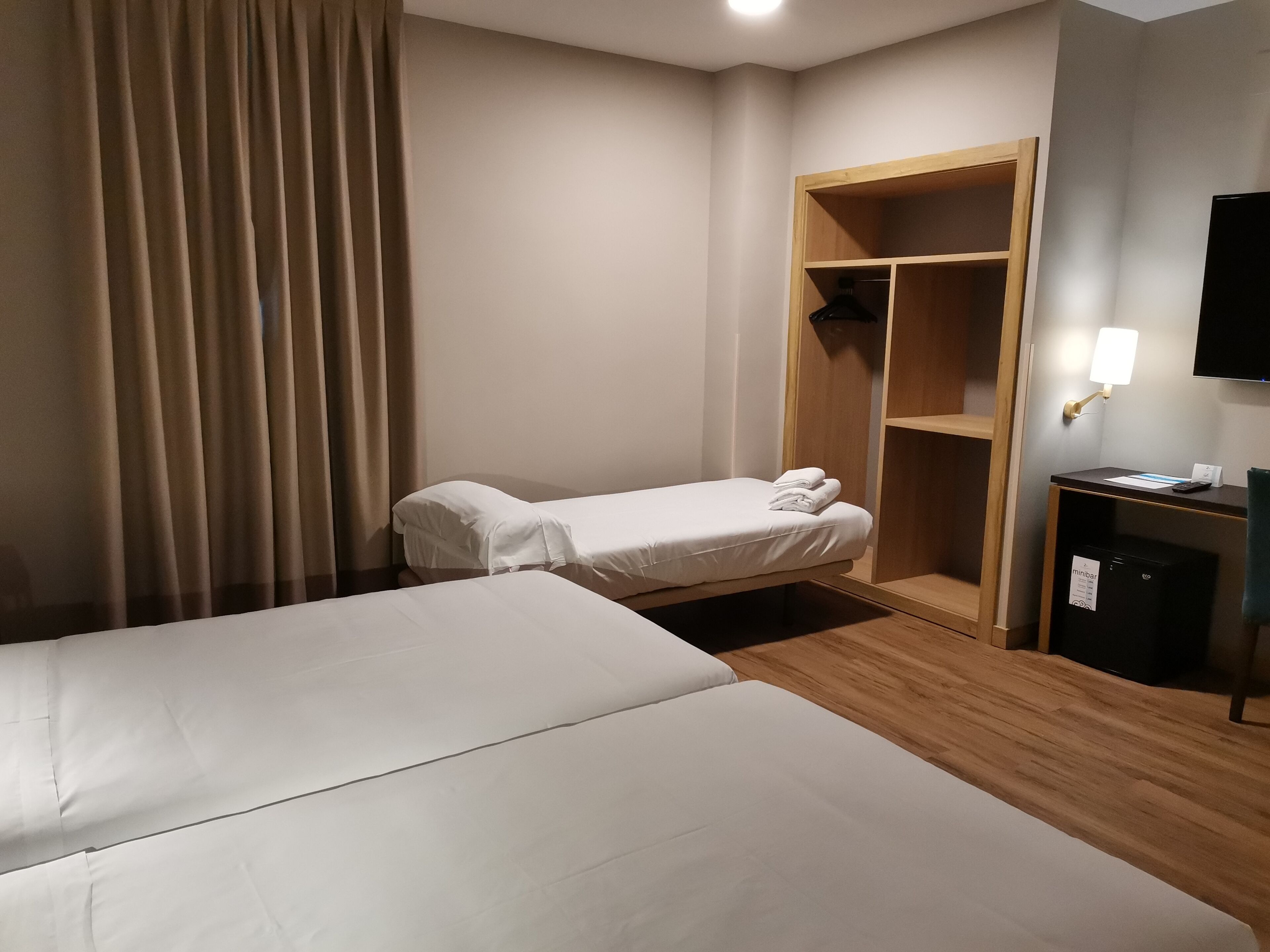 A hotel room with two beds, a wardrobe, a desk with a lamp, and a television.