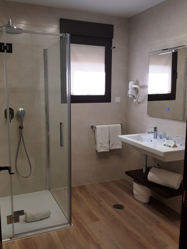 Un baño moderno con cabina de ducha acristalada, un lavabo blanco con espejo y una ventana con persiana oscura.