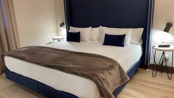 Una cama bien hecha con un cabecero oscuro, ropa de cama blanca y una manta marrón. Hay una mesita de noche con una lámpara y un teléfono. La habitación tiene suelo de madera y una pared de color claro.