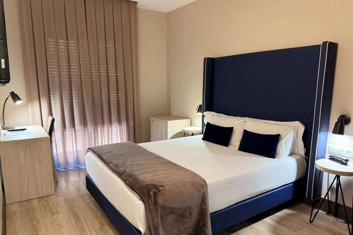 A hotel room with a large bed, a desk, a chair, a small table, and a window with curtains.