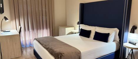 A hotel room with a large bed, a desk with a lamp, a small table, and a window with curtains.