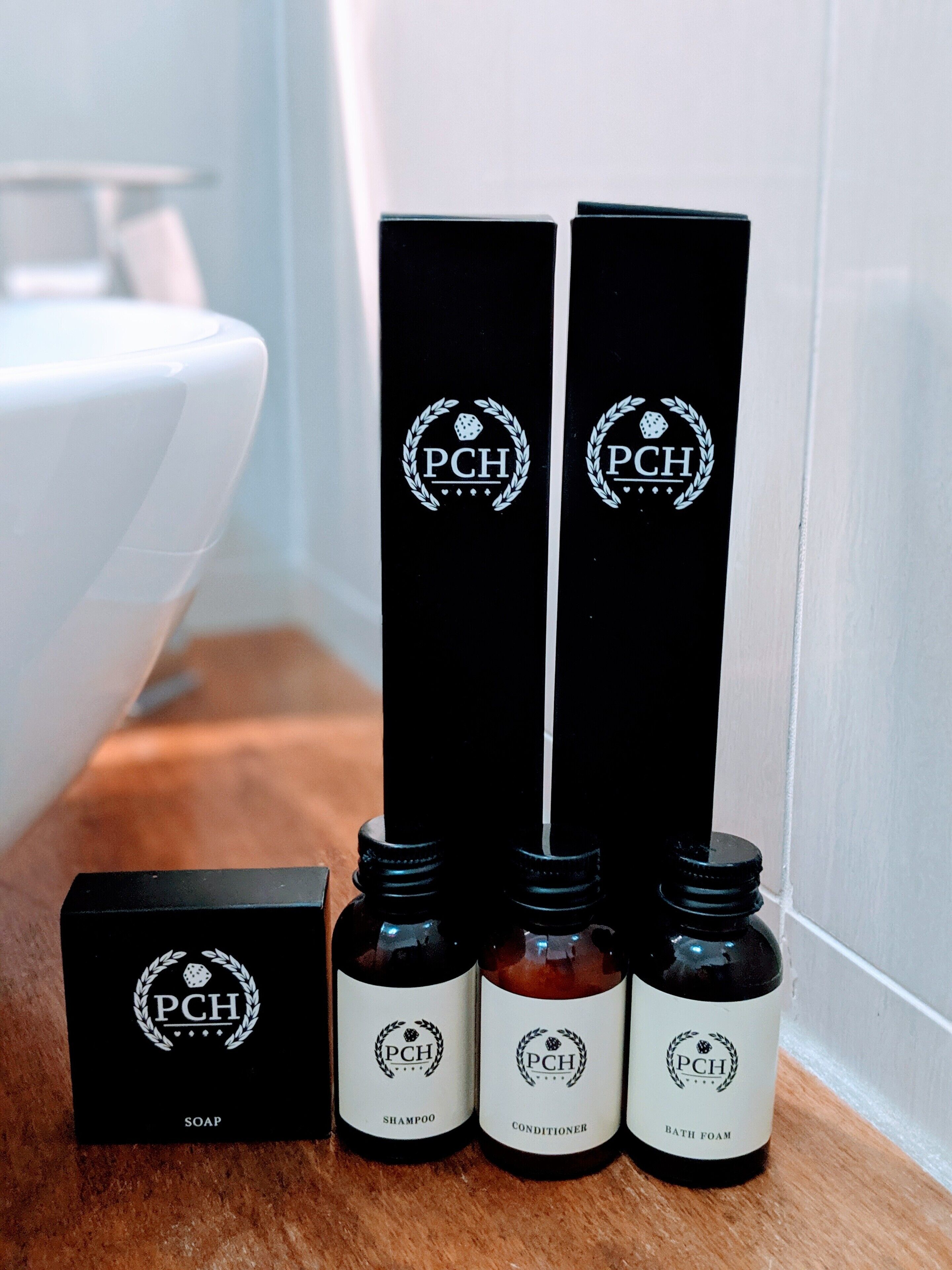 Ein Badezimmer mit einer weißen Badewanne, einem schwarzen Seifenspender und drei Flaschen mit Produkten der Marke PCH (Shampoo, Conditioner und Badeschaum) auf einer Holzfläche.