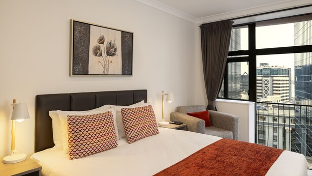 Une chambre d’hôtel avec un grand lit, deux coussins à motifs, une table de chevet avec une lampe, une chaise et une vue sur la ville à travers de grandes fenêtres.
