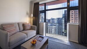 Une chambre d’hôtel moderne avec un canapé, une table basse sur laquelle se trouvent des fruits, et une grande fenêtre donnant sur la ville.