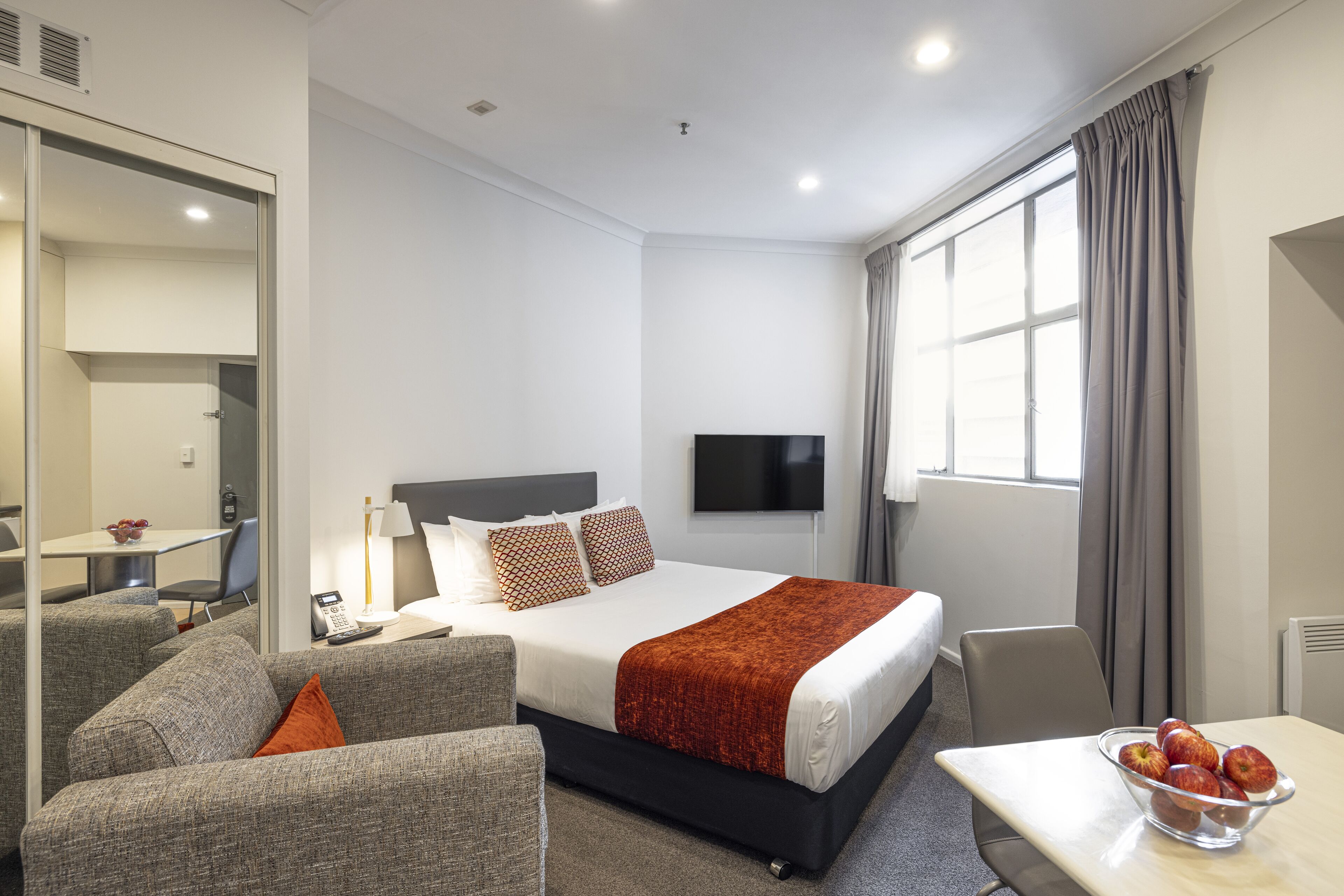 A hotel room with a bed, a TV, a desk with a bowl of fruit, and a sofa.