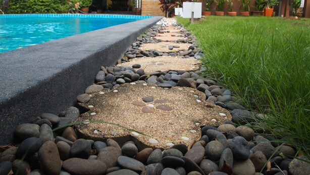 Un sendero conduce a una piscina con borde de piedra y piedras, rodeada de follaje y plantas en macetas.