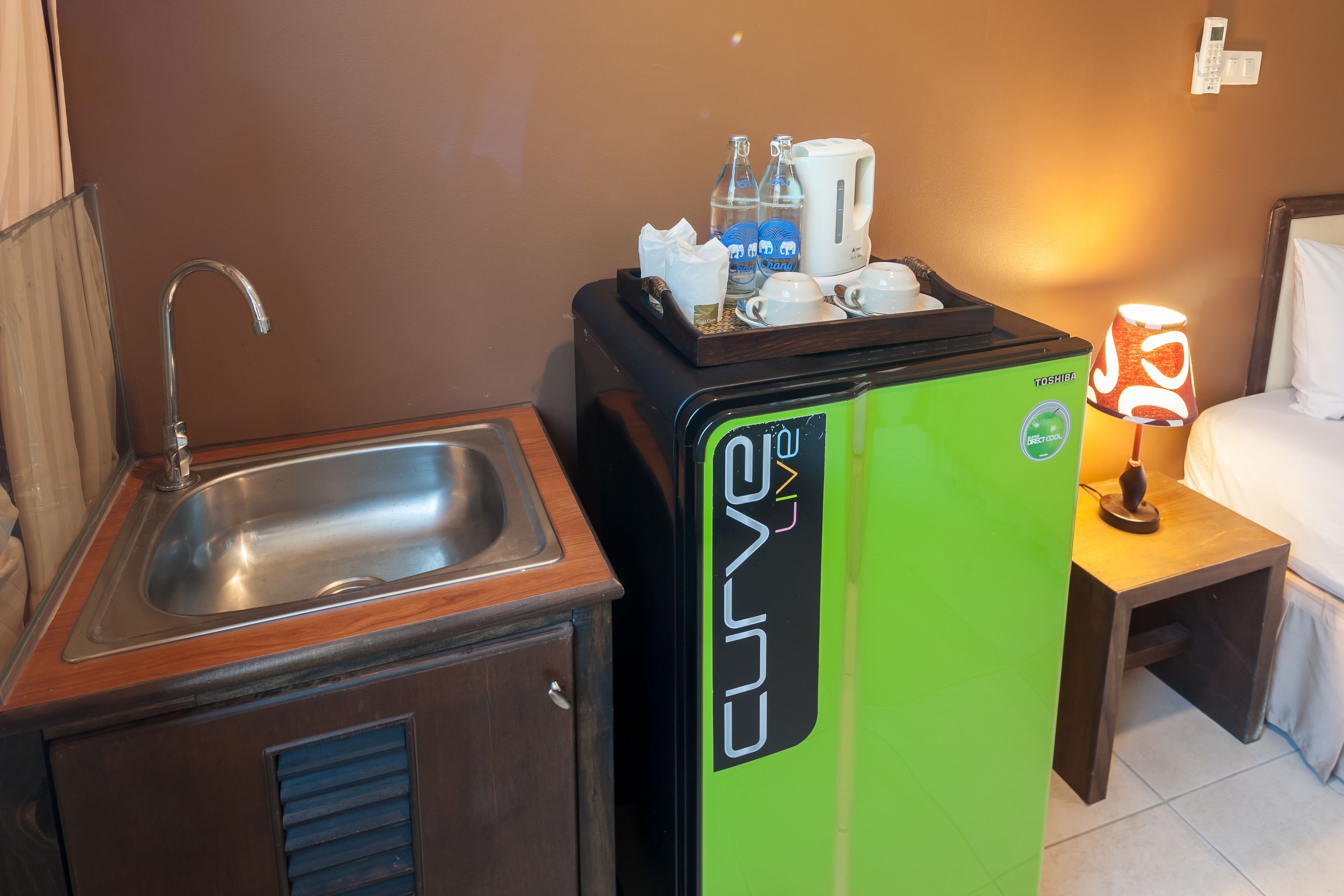 A compact kitchenette with a sink, a small cabinet, a green refrigerator, and a bedside table with a lamp.