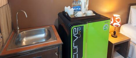 A compact kitchenette with a sink, a small cabinet, a green refrigerator, and a bedside table with a lamp.