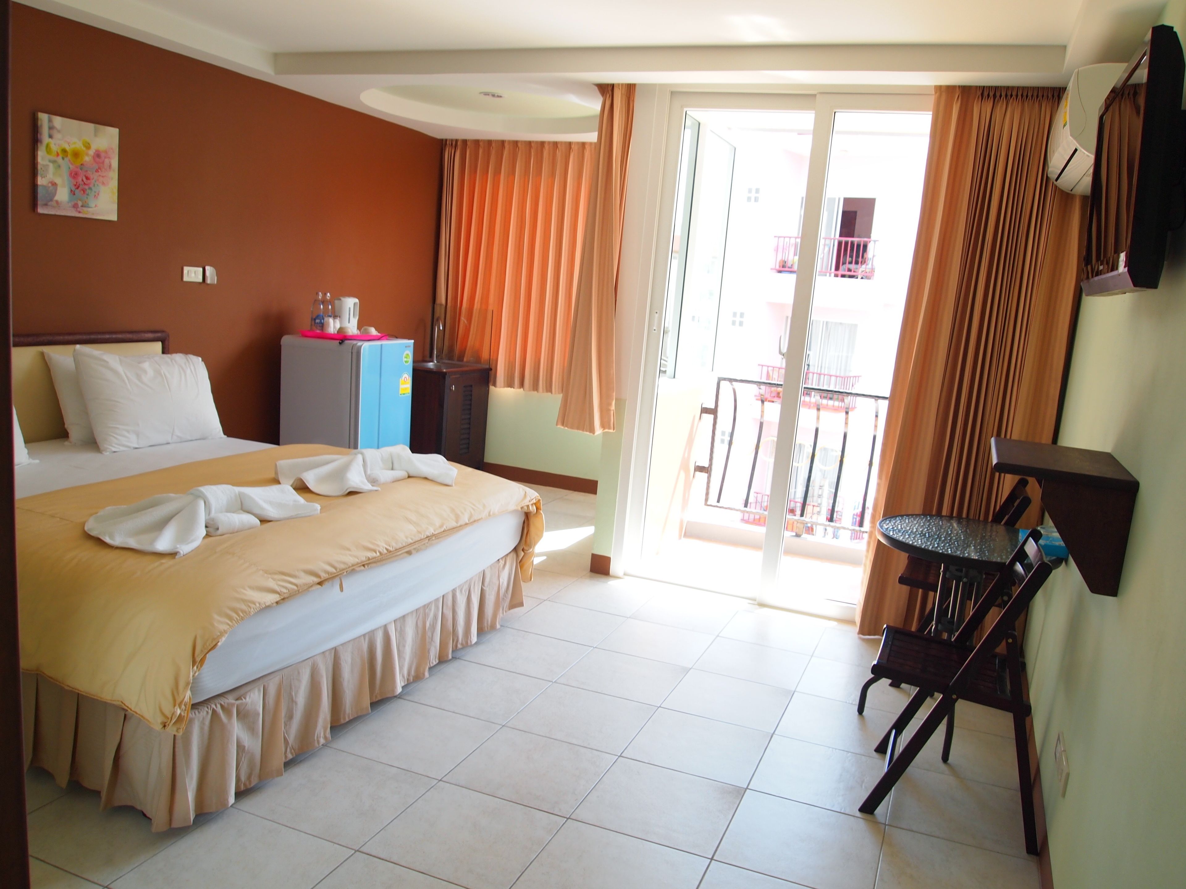 A hotel room with a bed, a desk, a chair, a small table, a refrigerator, and a sliding glass door leading to an outdoor balcony.