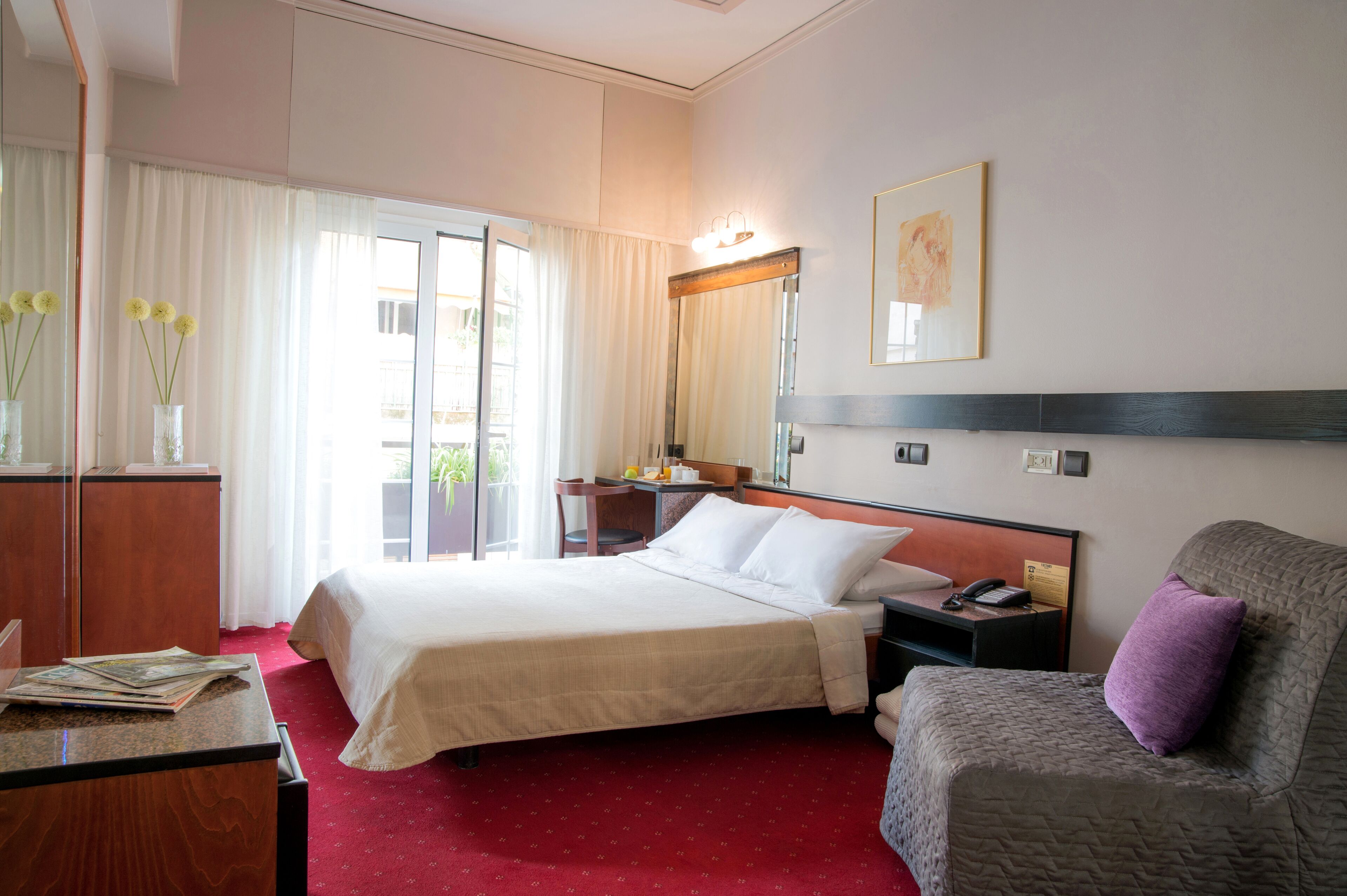 Habitación de hotel con una cama grande, un escritorio, una silla, una mesita, una mesita de noche, una lámpara, un espejo y una ventana con cortinas.