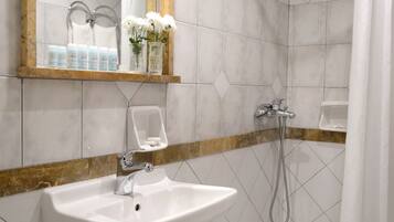 A bathroom with a sink, mirror, shower, and toiletries on the shelf.