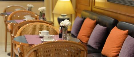 A dining area with wicker chairs, a table with a purple placemat, white flowers, and a sofa with orange and purple cushions.