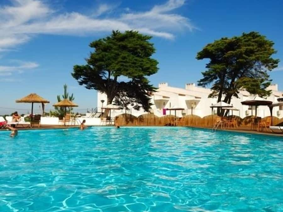 A swimming pool with lounge chairs and umbrellas, surrounded by white buildings and trees.
