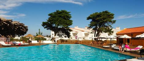 A swimming pool with wooden deck and lounge chairs, surrounded by white buildings and trees.