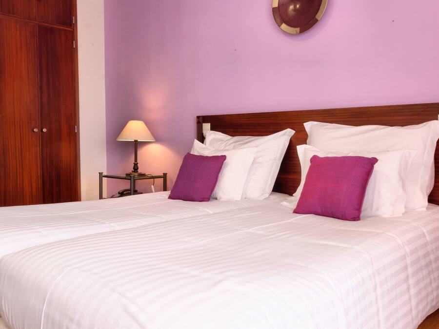A double bed with white bedding and purple pillows, a wooden headboard, a nightstand with a lamp, and a wooden wardrobe.