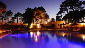 Area piscina illuminata di notte, con poltrone relax e un edificio sullo sfondo.