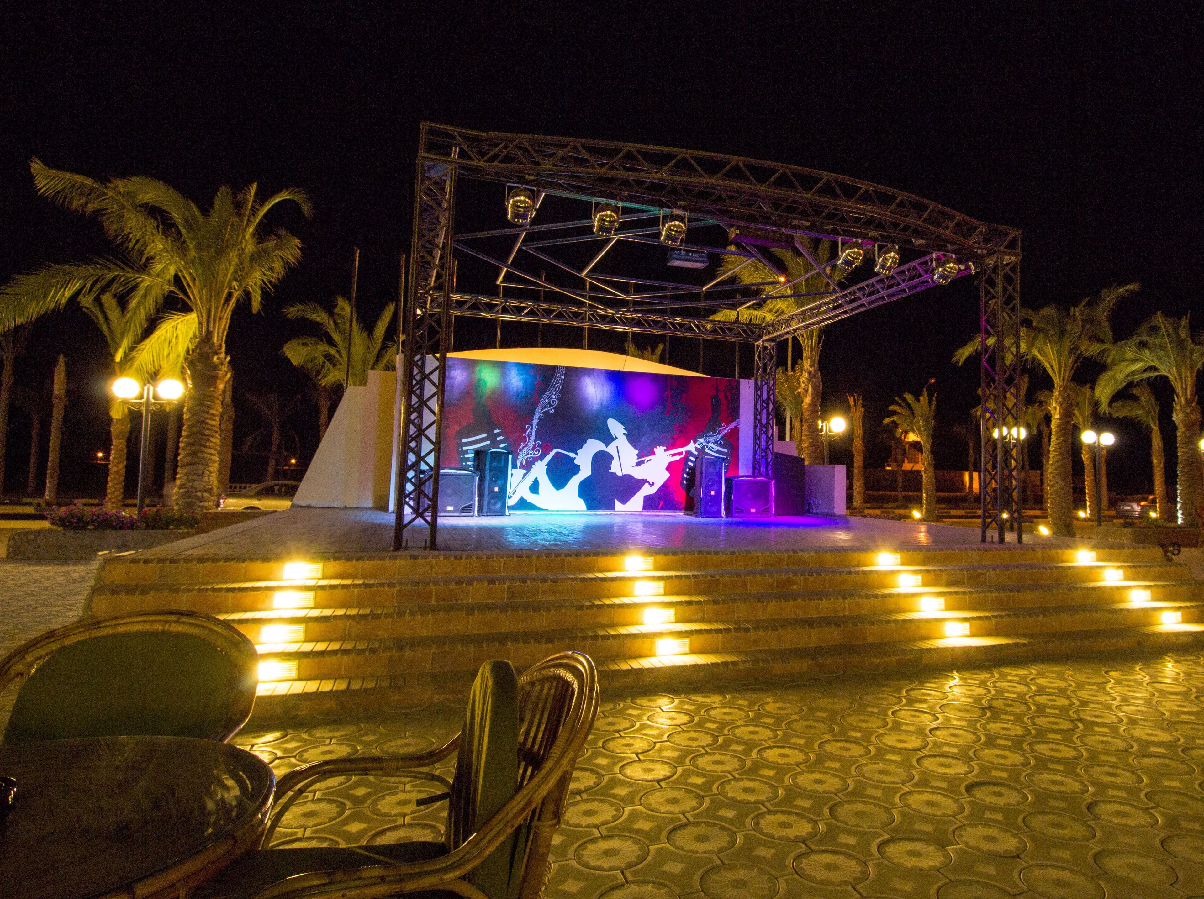 An outdoor stage with a band performing, surrounded by palm trees and a well-lit seating area.