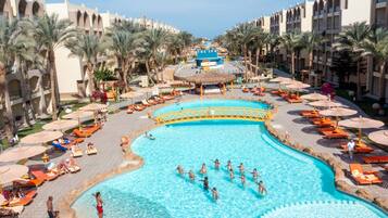 A large pool surrounded by sun loungers and umbrellas, with palm trees and a hotel building in the background.