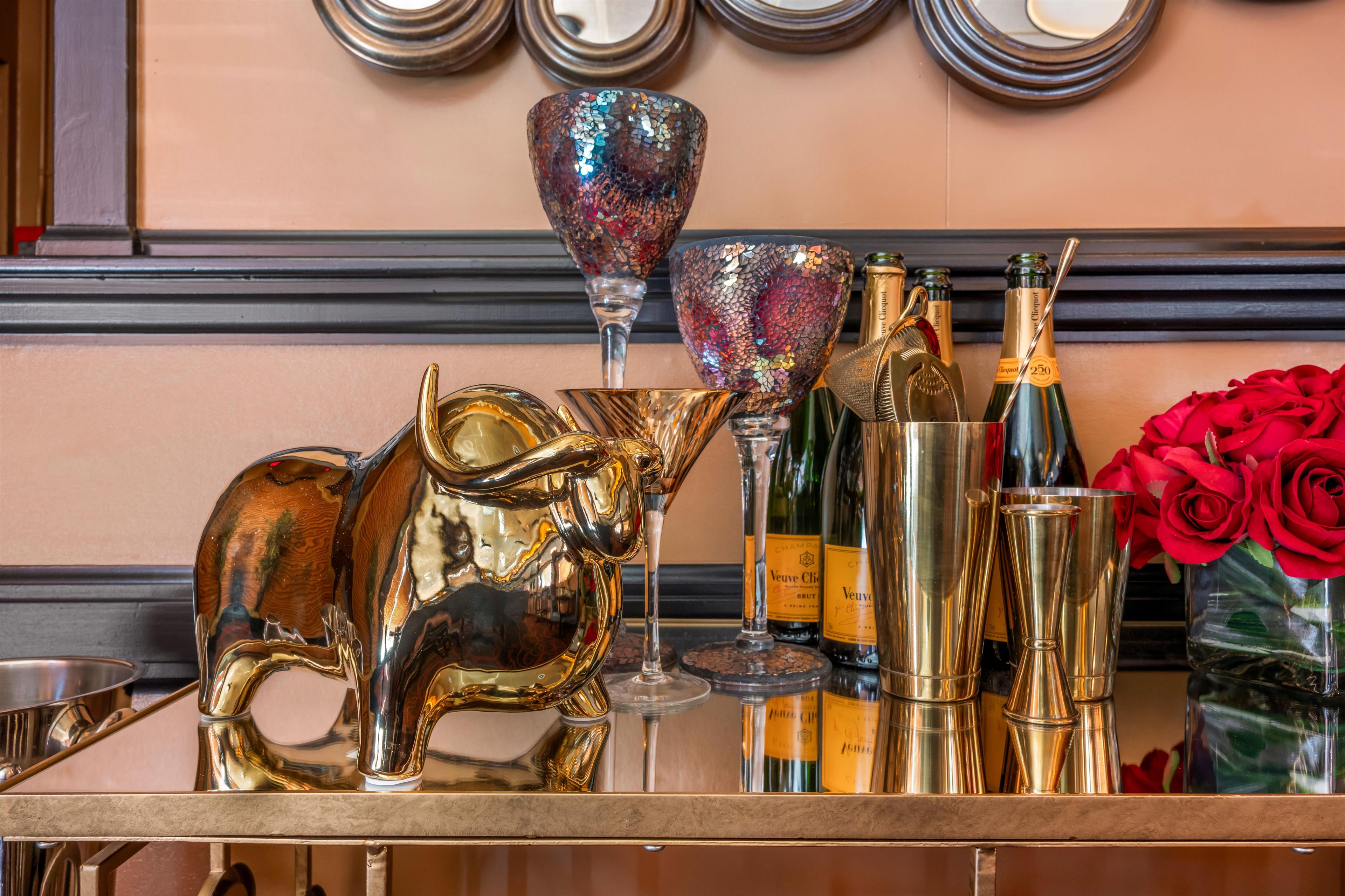 A bar area with a decorative bull sculpture, a glass with a mosaic pattern, champagne bottles, and a vase of red roses.