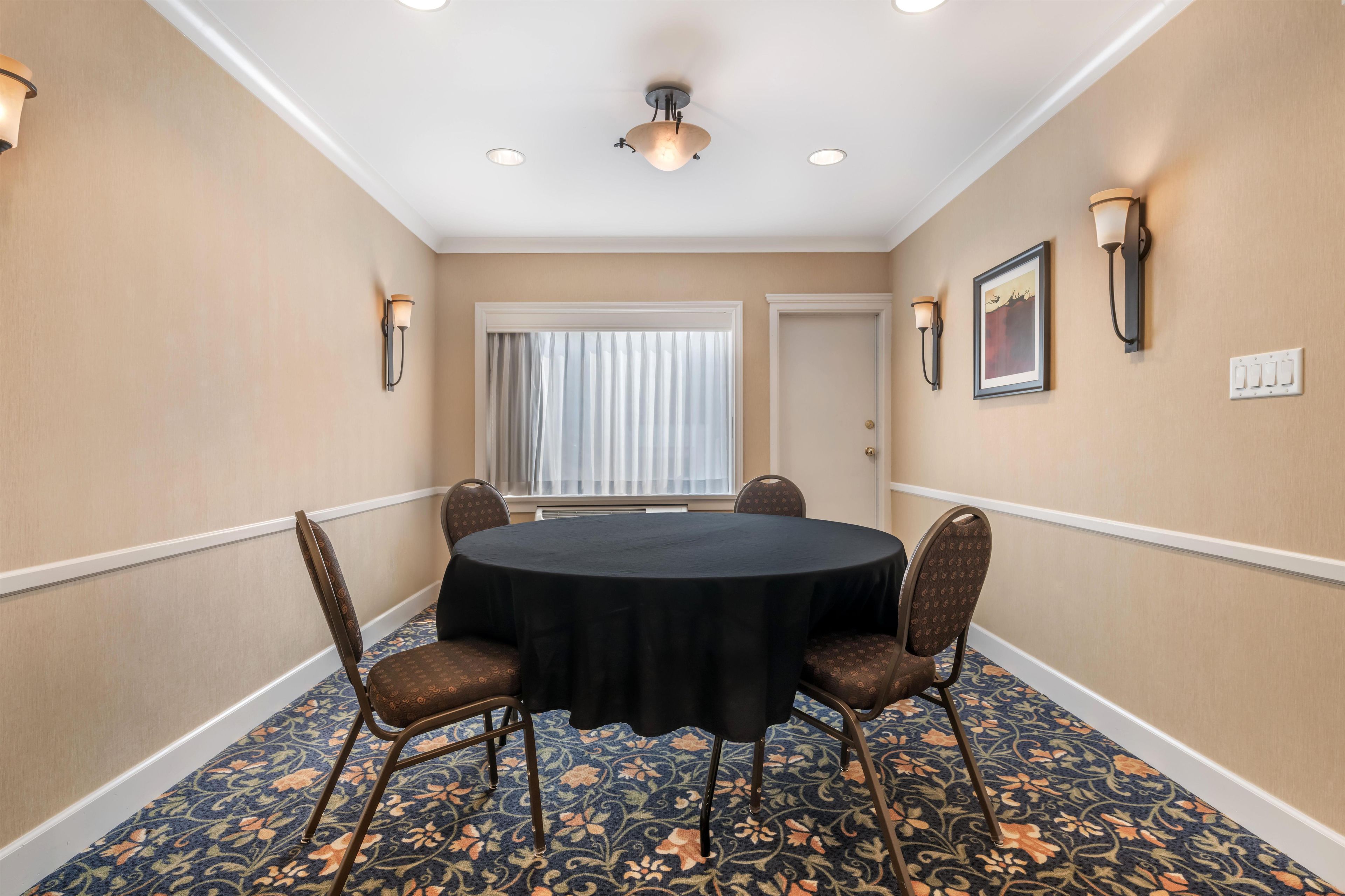 A conference room with a round table, four chairs, a window with curtains, wall sconces, and a framed picture.
