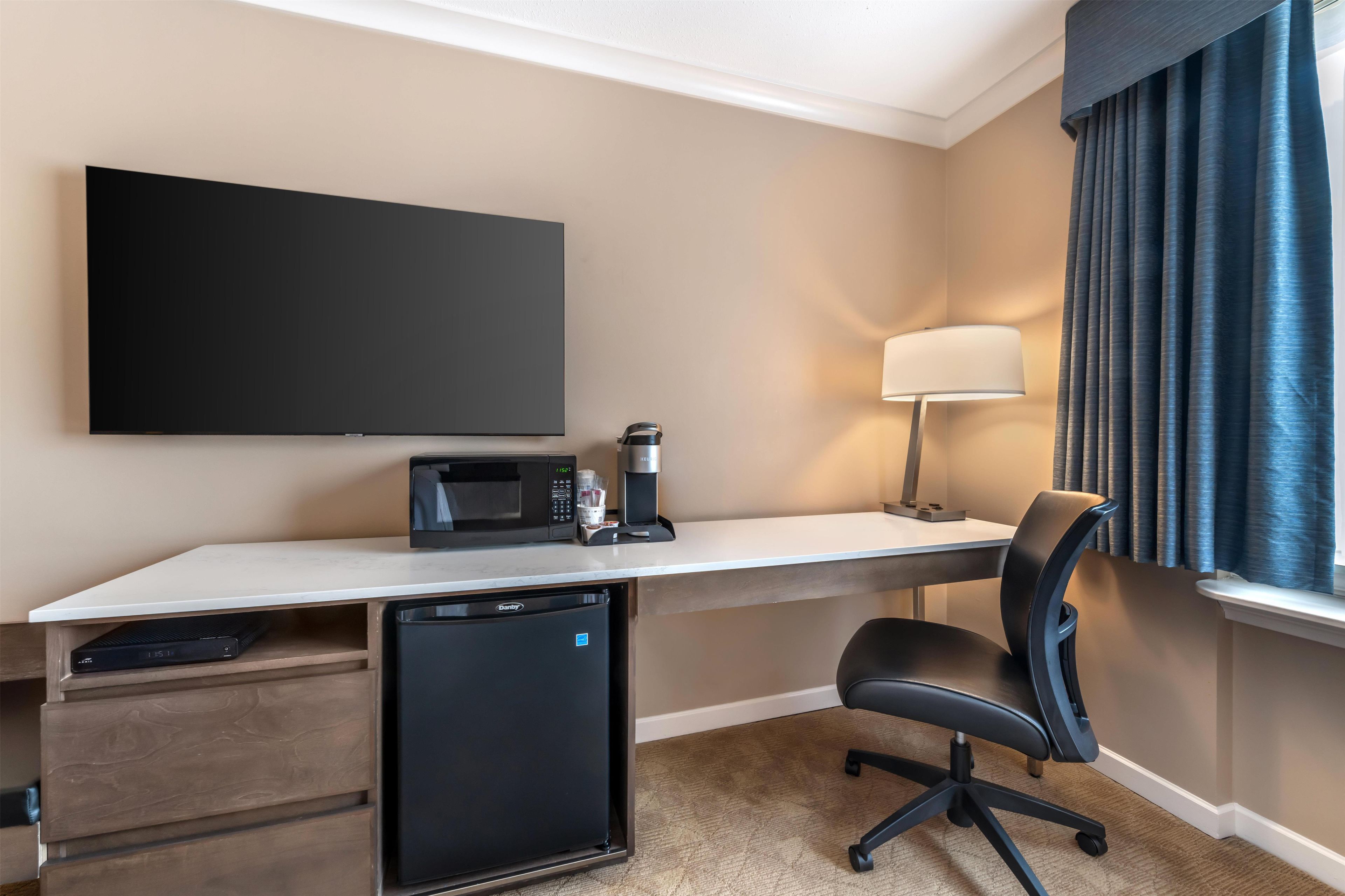 Habitación de hotel con televisión de pantalla plana, cafetera, microondas, refrigerador pequeño, escritorio, silla y ventana con cortinas.