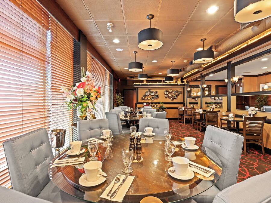 A dining area with a round table set for a meal, surrounded by chairs, and a large window with blinds.