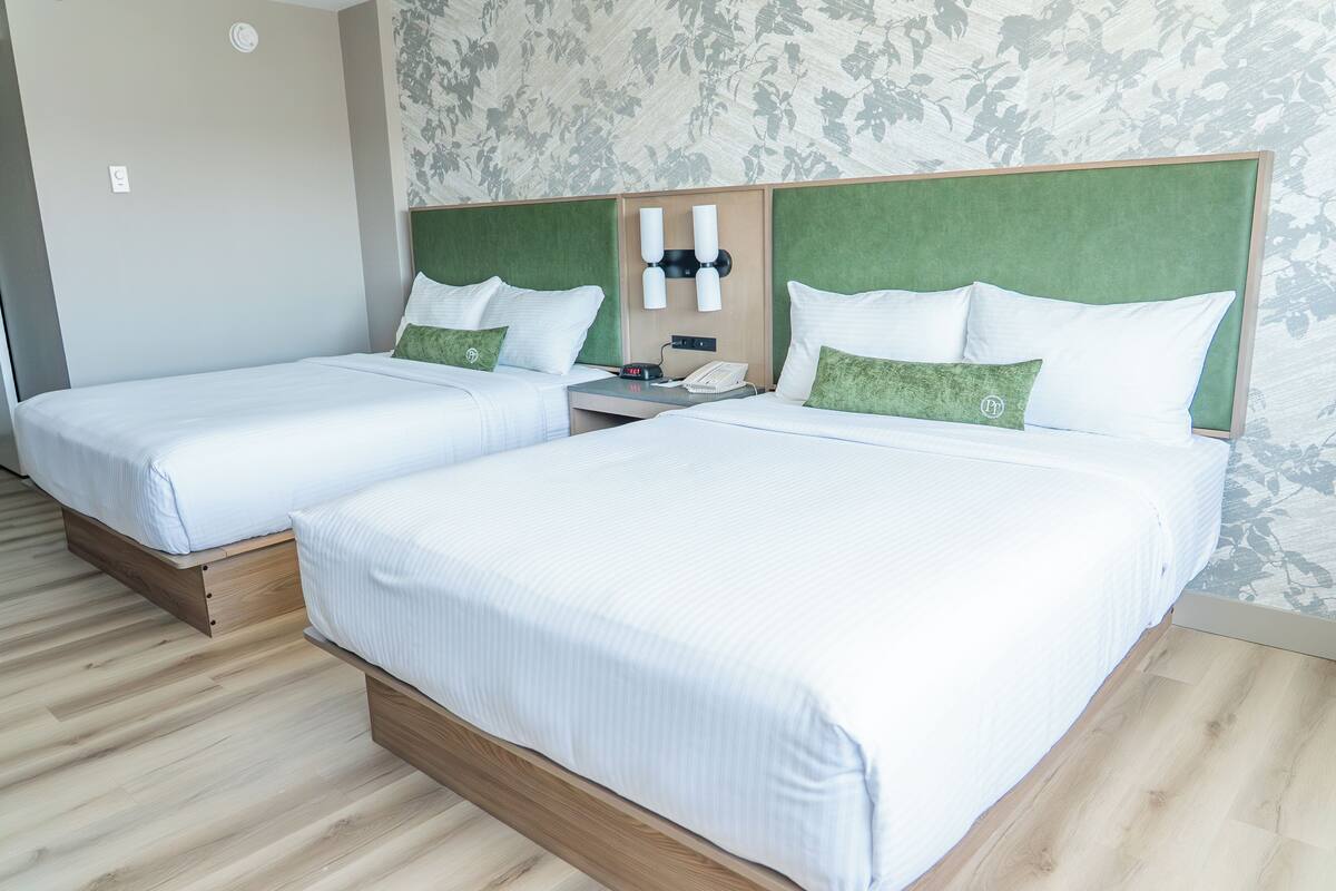 A hotel room with two single beds, wooden headboards, a nightstand with a phone and alarm clock, and a floral-patterned wall.