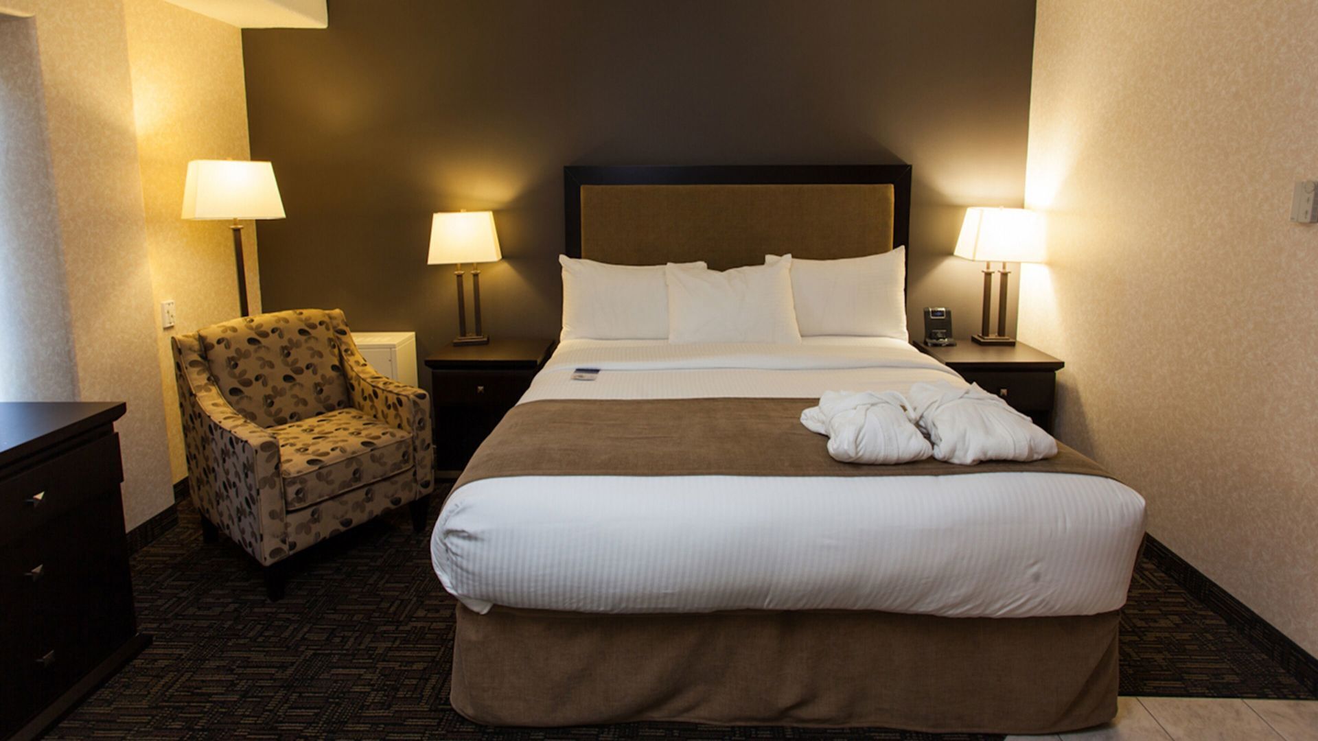 A hotel room with a large bed, two bedside lamps, a patterned armchair, and a nightstand with a phone.