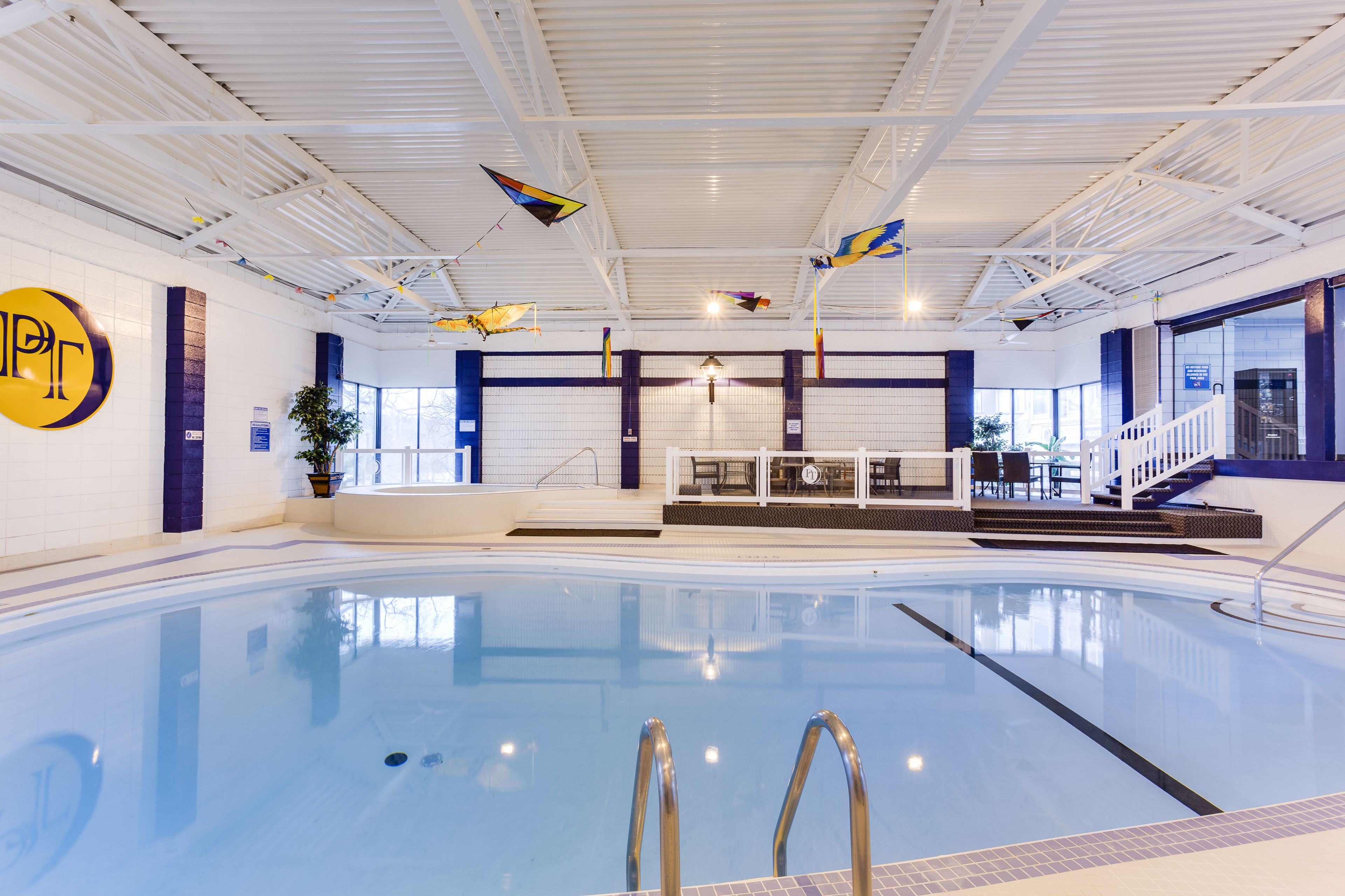 An indoor swimming pool with clear water, a visible logo on the wall, and a staircase leading to a seating area.